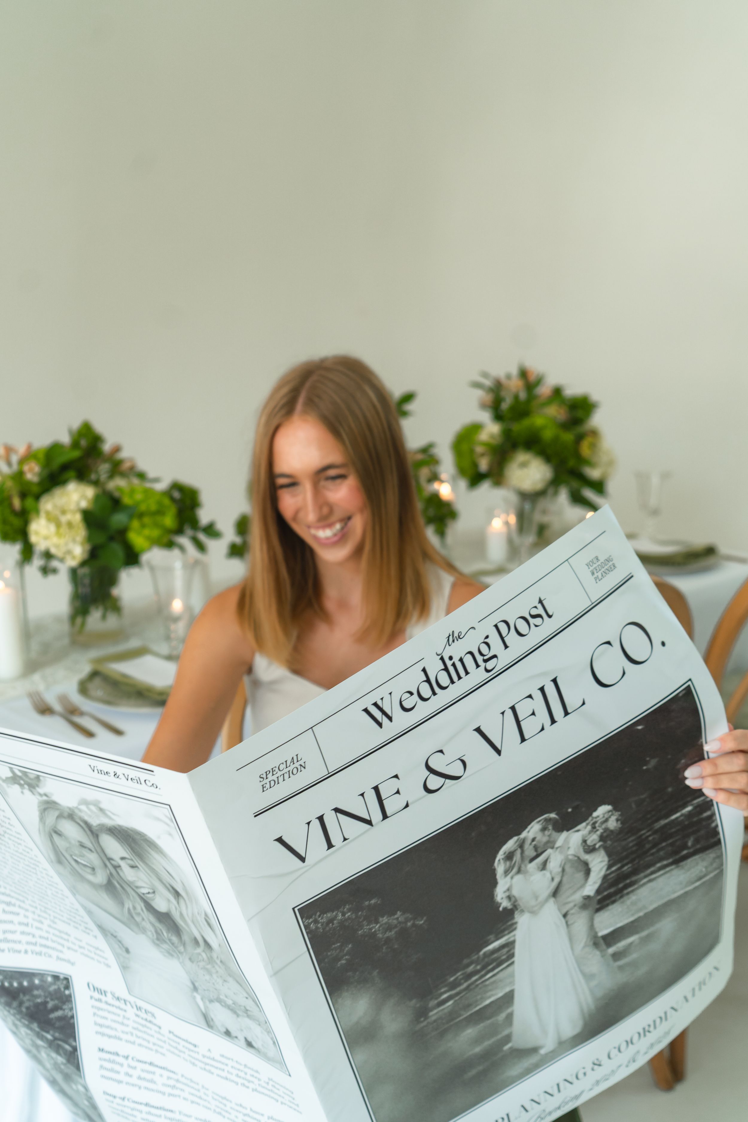 Smiling bride reads Vine & Veil Co. Wedding Post newspaper at elegant floral reception table.