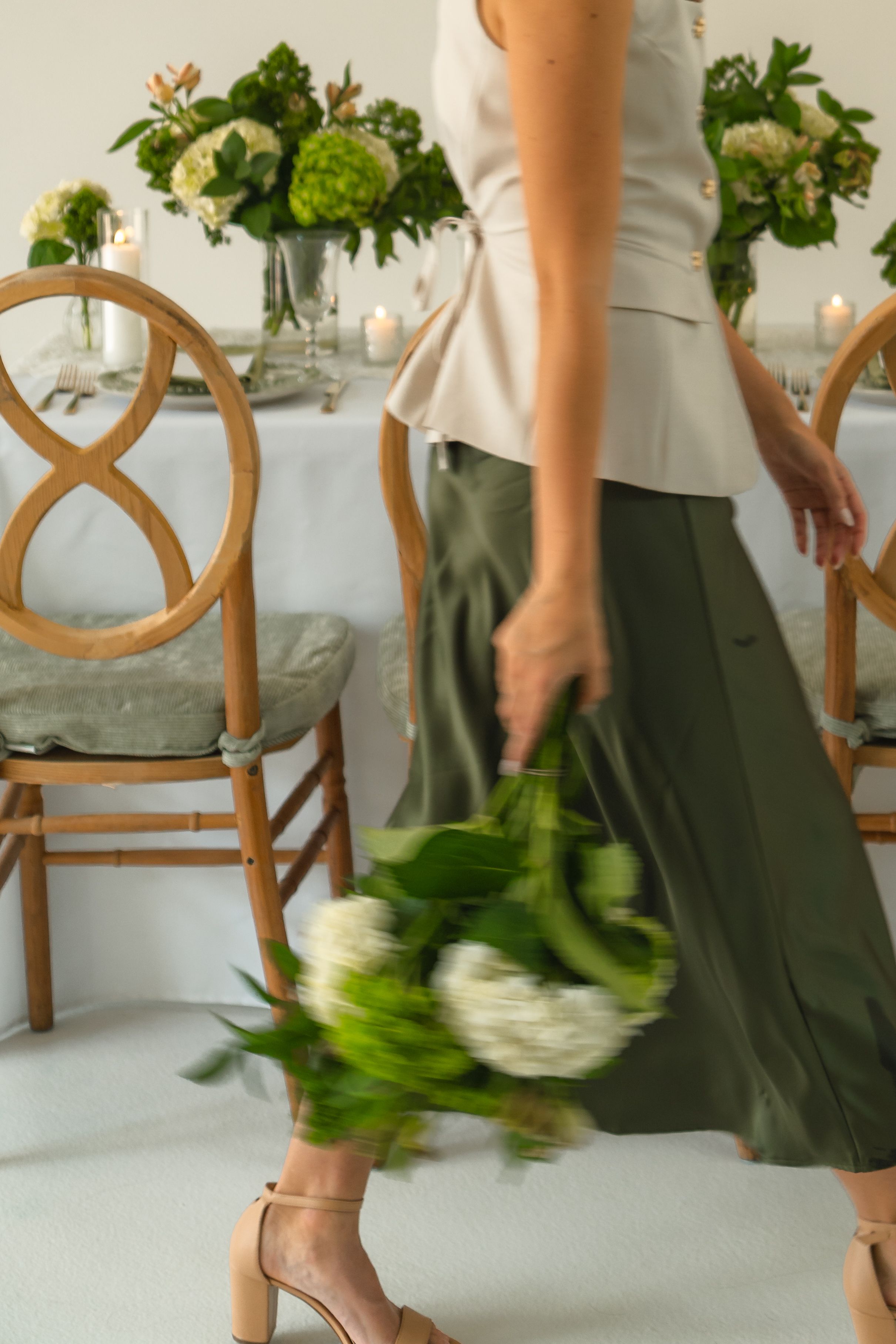 Woman in green skirt holding white bouquet walks past elegant table setting with floral arrangements and wooden chairs.