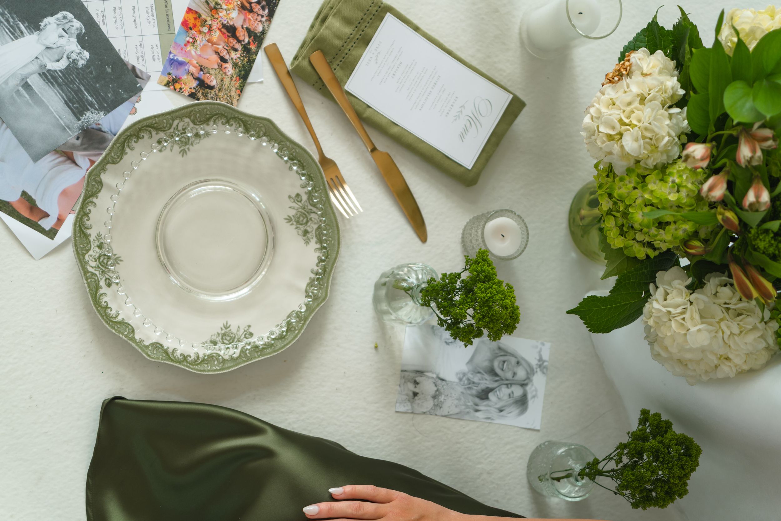 Elegant table setting with green floral plate, gold cutlery, hydrangeas, and candles on white linen.