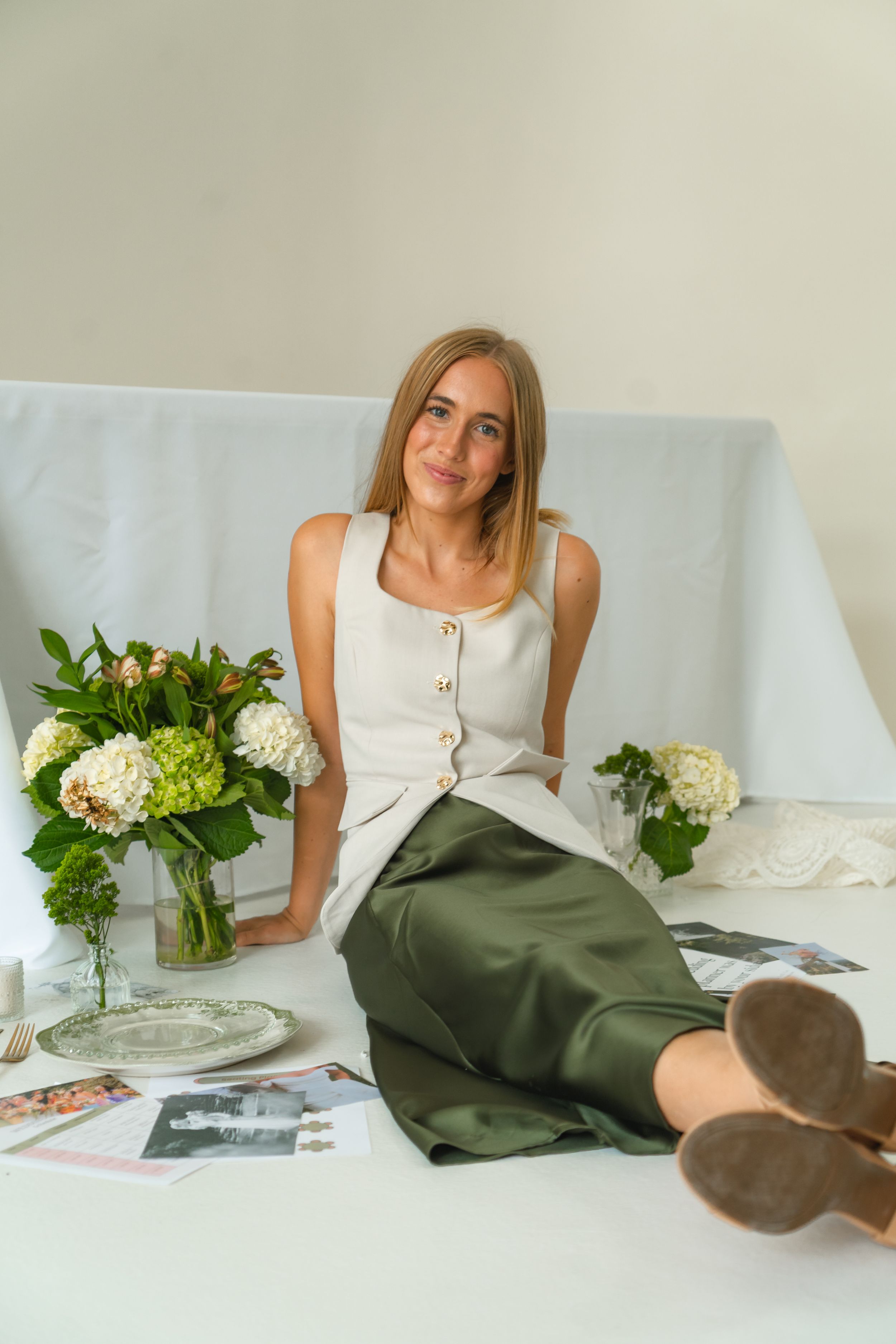 Woman in white top and green skirt sitting on floor with hydrangea flowers and magazines in bright studio.