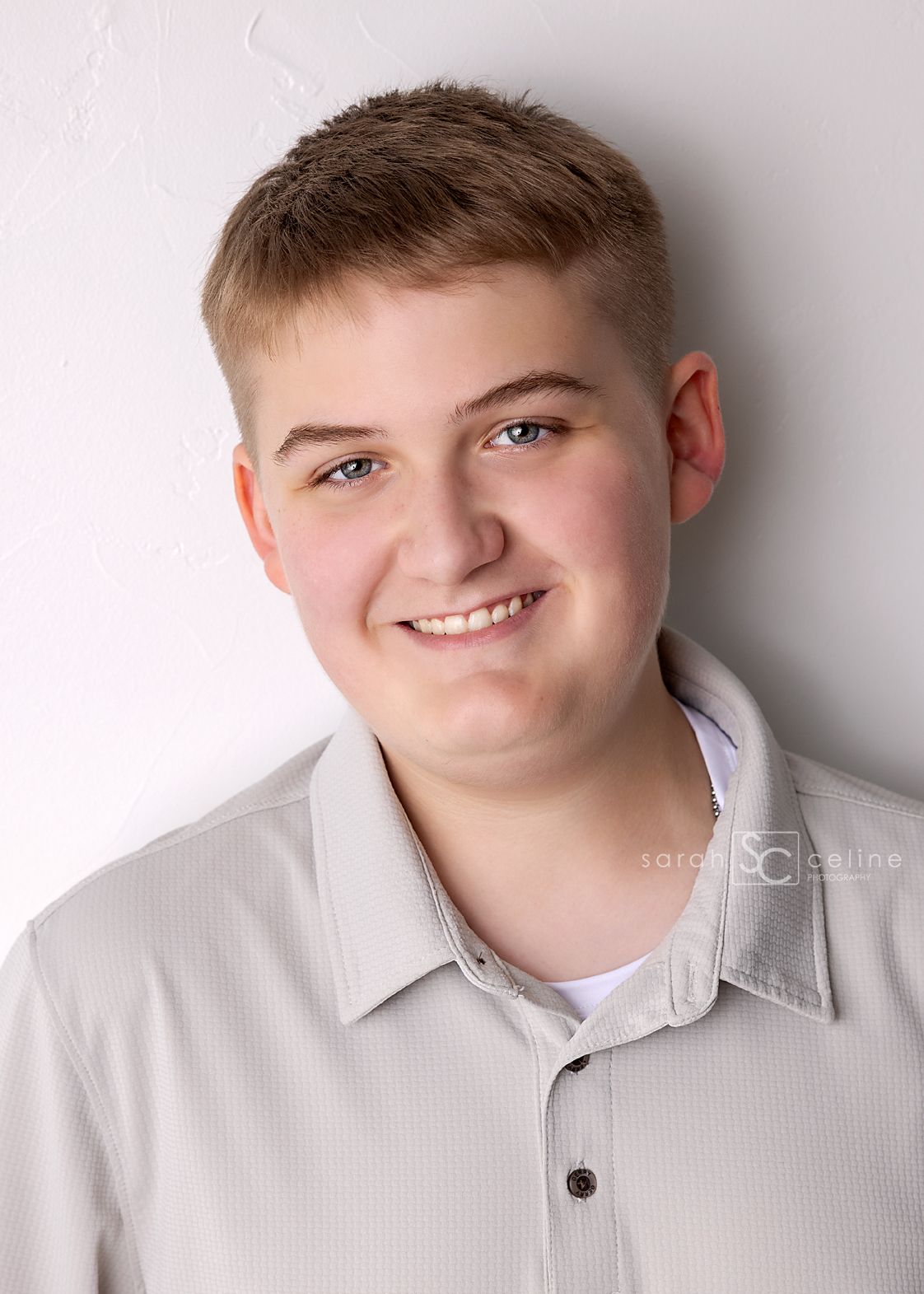 Smiling teenage boy with short blonde hair wearing a light gray polo shirt against a white background.