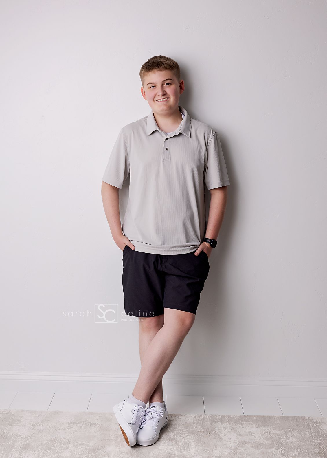 Smiling teenage boy in gray polo shirt and black shorts leaning against a white wall in a studio portrait.