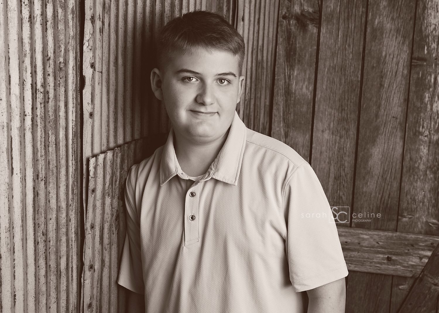 Young man in polo shirt smiling, leaning against rustic wooden wall in sepia-toned portrait photo.
