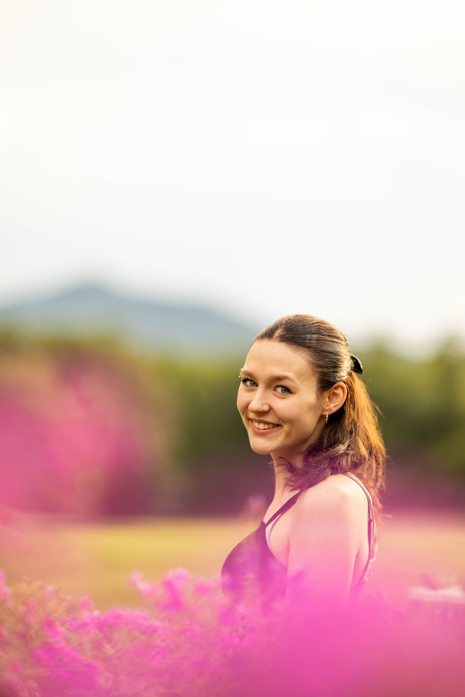 Young woman smiling in a vibrant pink flower field with mountains and green trees in the background.