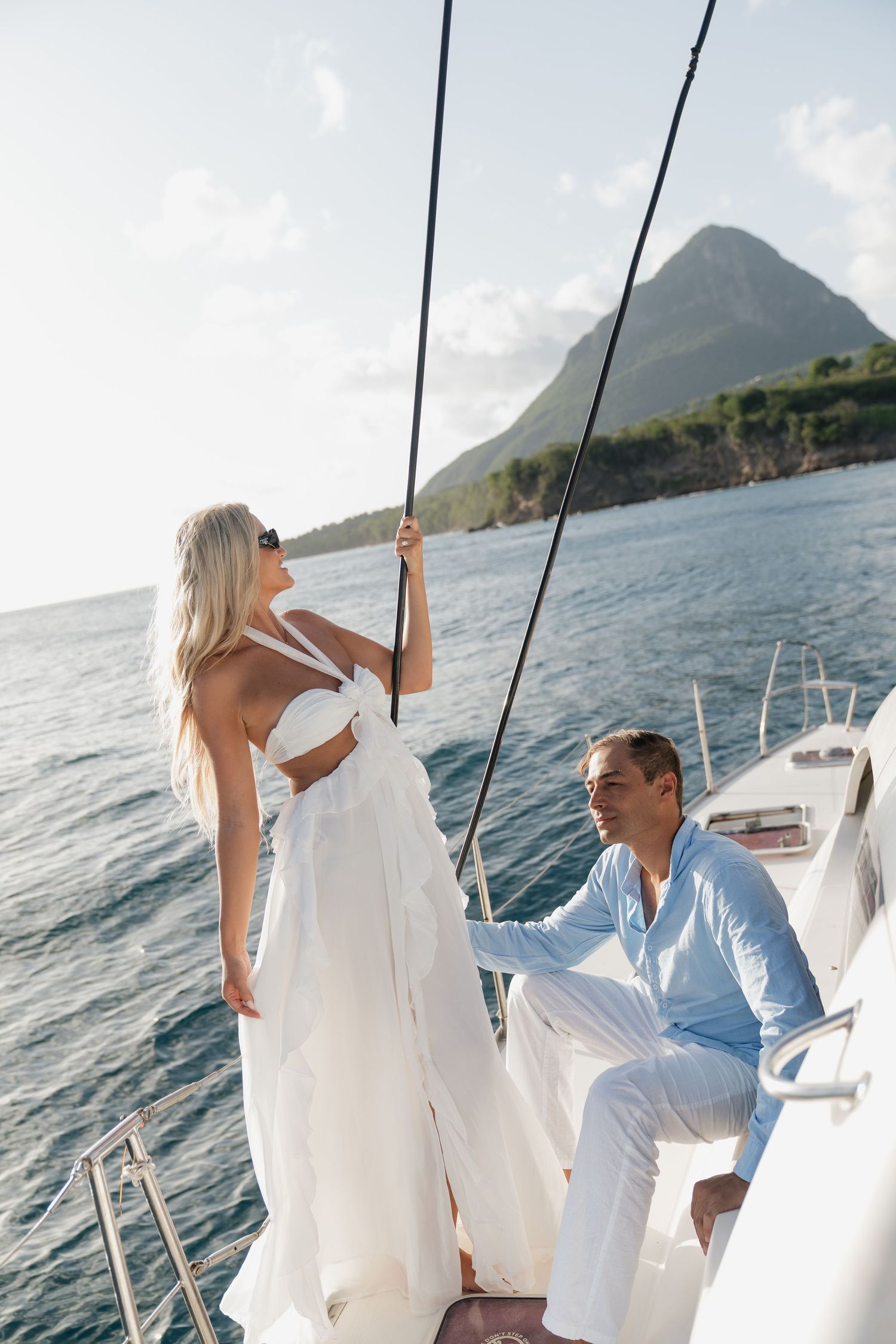 Elegant couple in white outfits on a luxury sailboat with a scenic mountain and ocean backdrop.