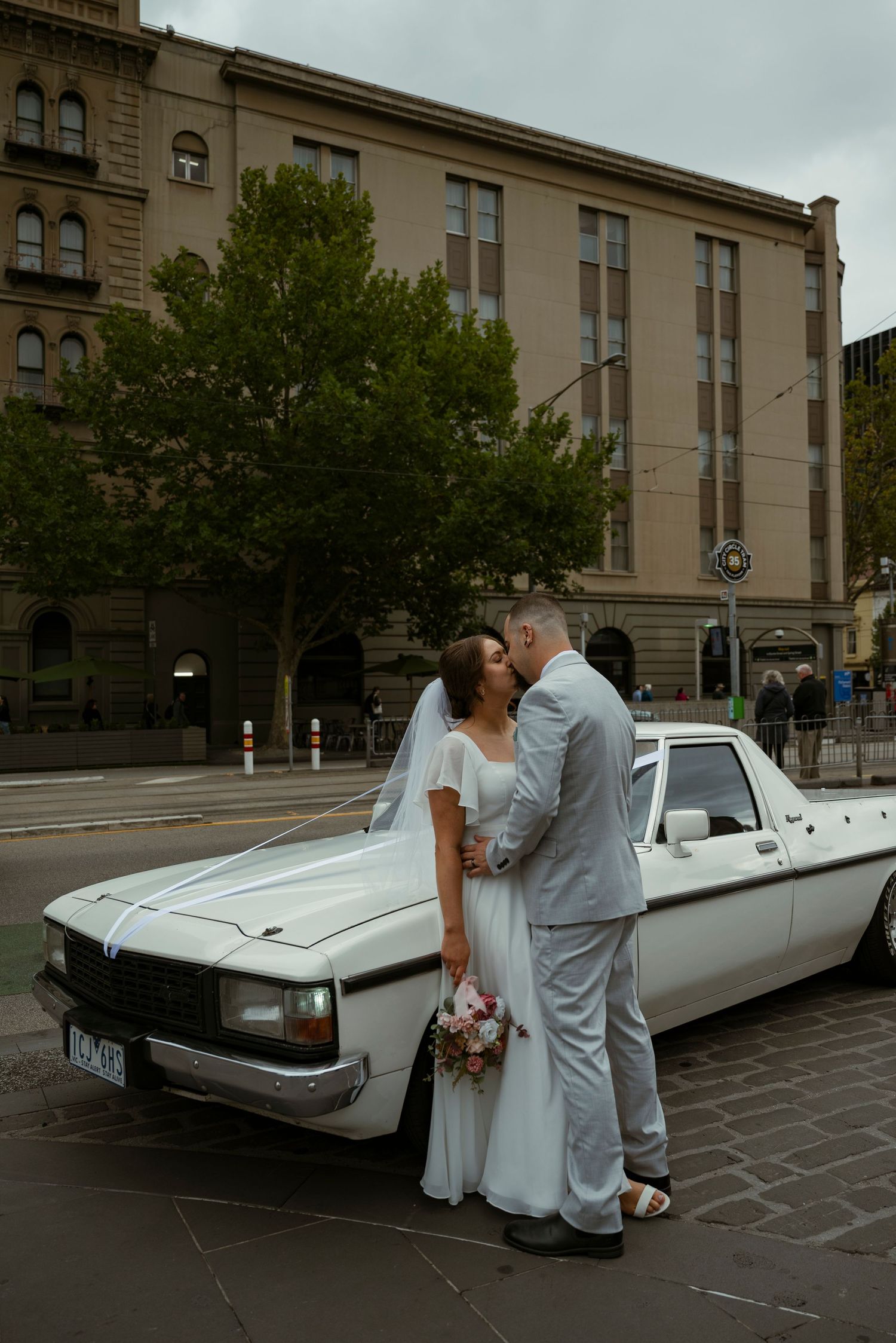 Bride and groom kissing beside a classic white car on a city street, bride holding a bouquet in a white wedding dress.