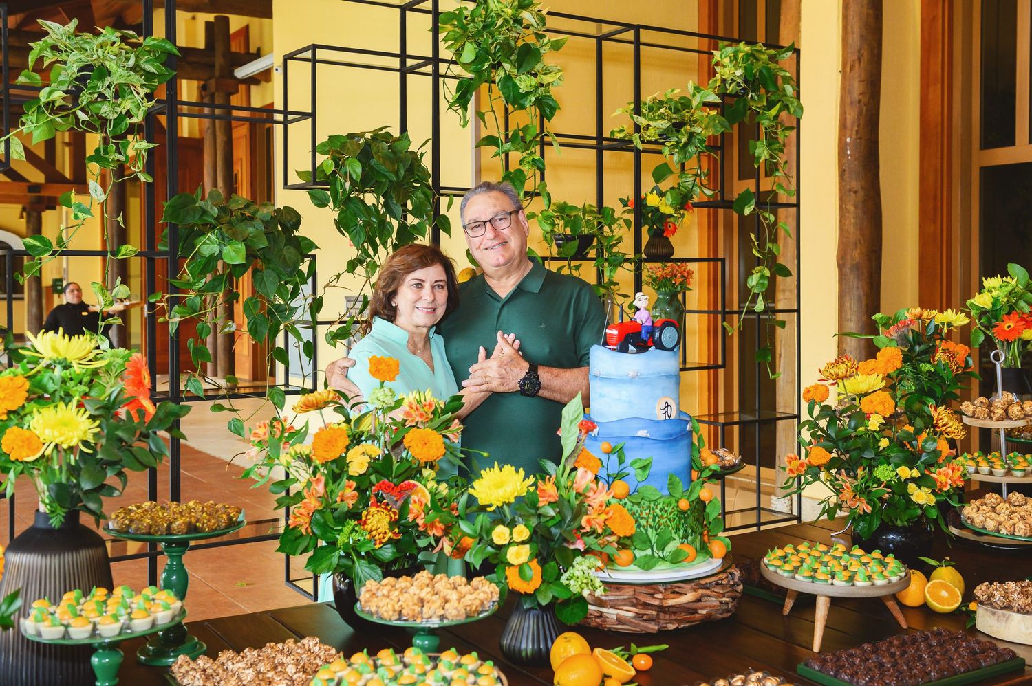Couple smiling together at a colorful party dessert table with floral arrangements, fruit decor, and a blue celebration cake.