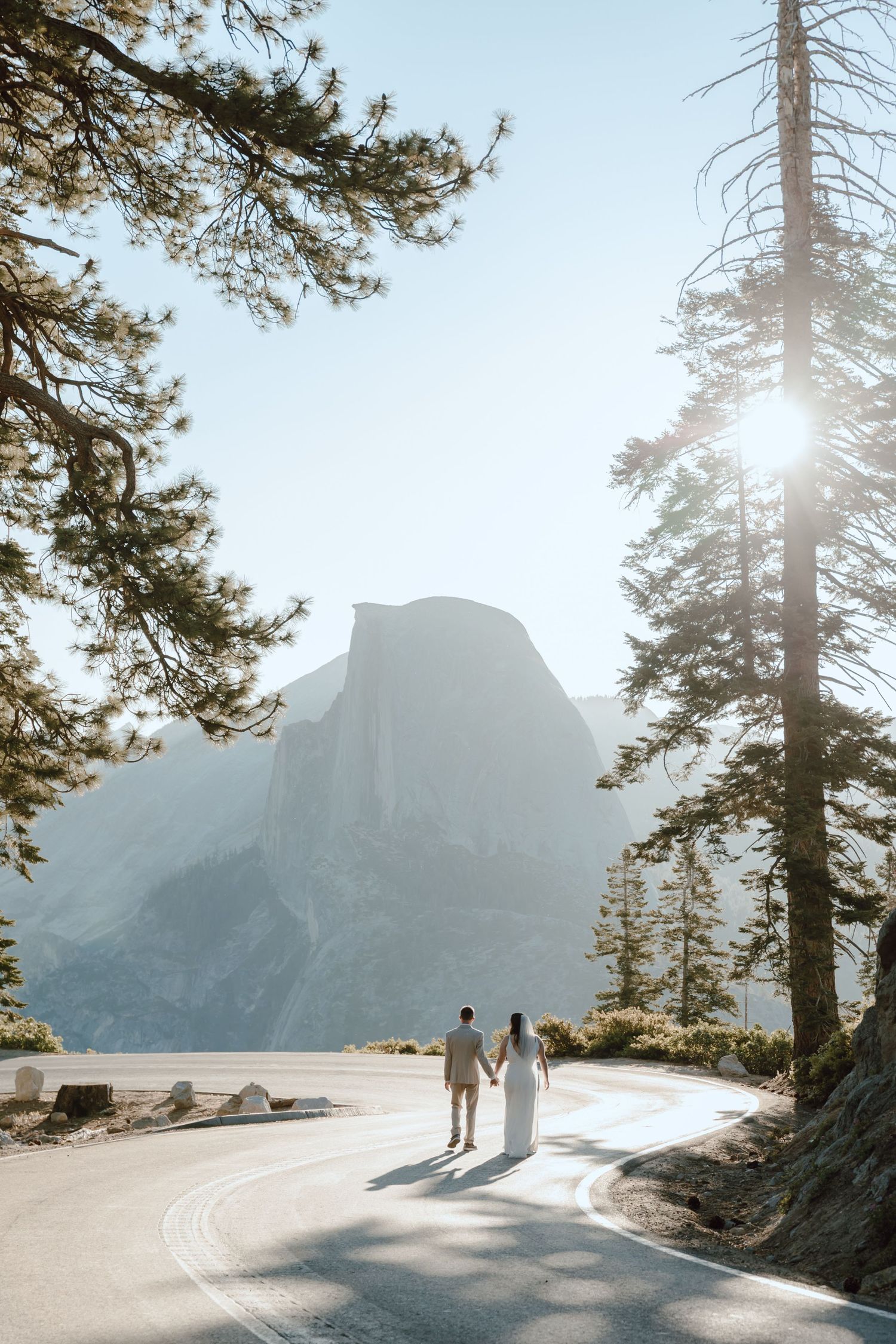 Couple walks hand-in-hand on a sunlit road in Yosemite with Half Dome in the background.