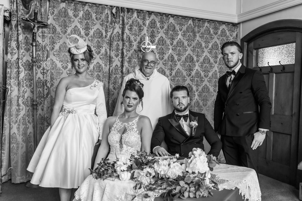 Black and white wedding photo of bride and groom signing register with three guests posing in elegant venue.