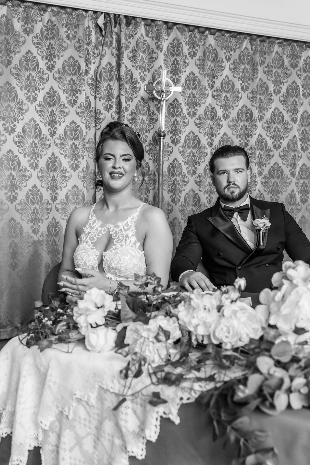 Black and white photo of bride and groom seated at wedding table with floral arrangements and ornate wallpaper backdrop.