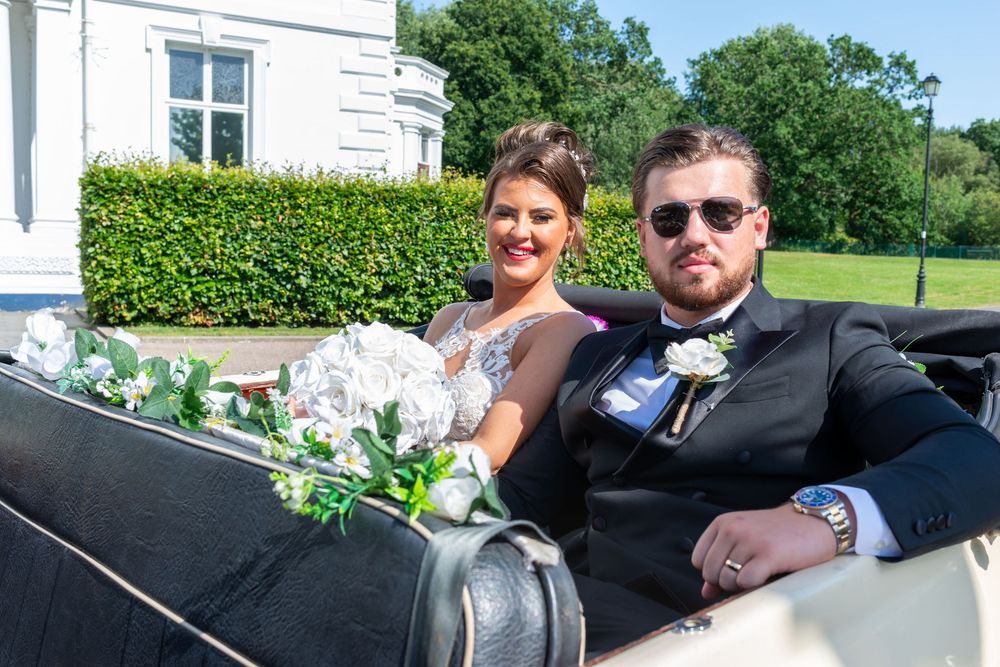 Bride and groom sitting in a vintage wedding car adorned with white flowers on a sunny day.