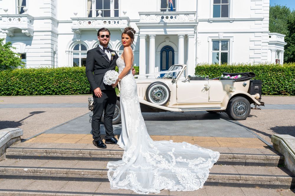 Bride and groom posing in front of a vintage cream convertible car outside an elegant white manor house.