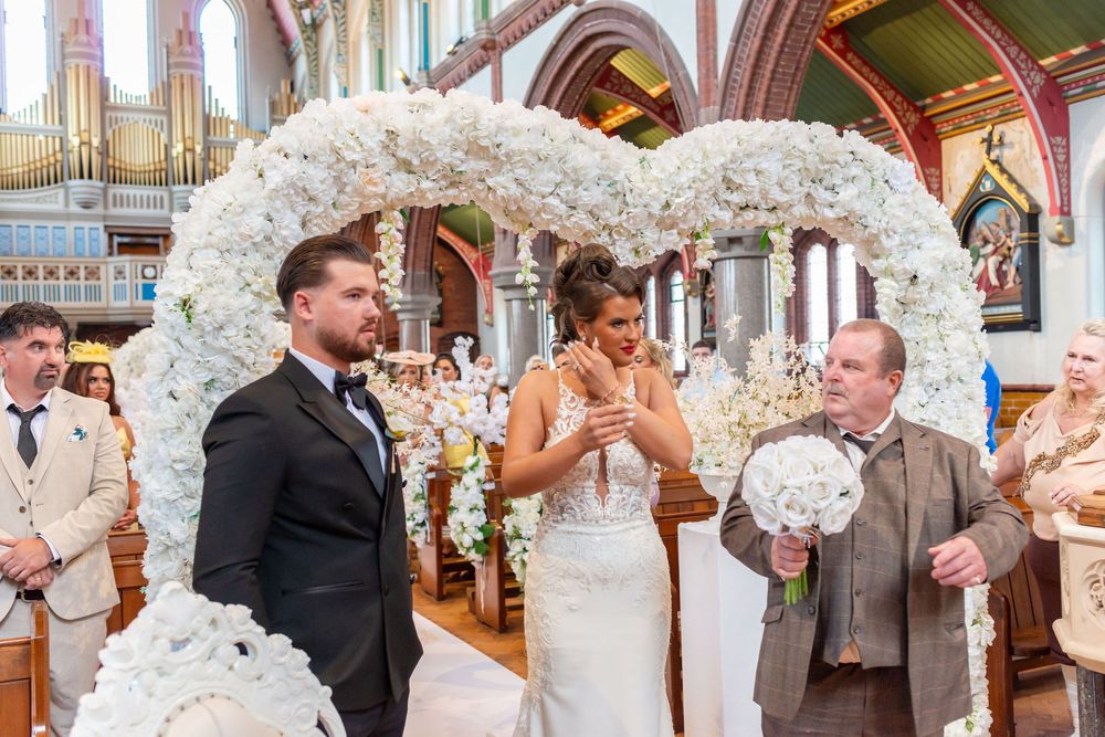 Bride and groom stand under white floral heart arch during wedding ceremony inside ornate church.
