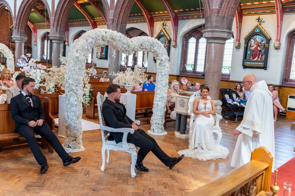Bride and groom seated at church altar under floral arch during wedding ceremony with priest and guests.