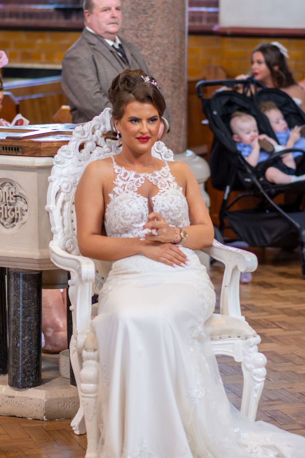 Bride in white lace wedding gown seated on ornate white chair during wedding ceremony in church.