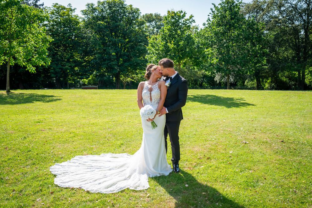 Bride and groom share a romantic kiss on a sunny green lawn surrounded by lush trees on their wedding day.