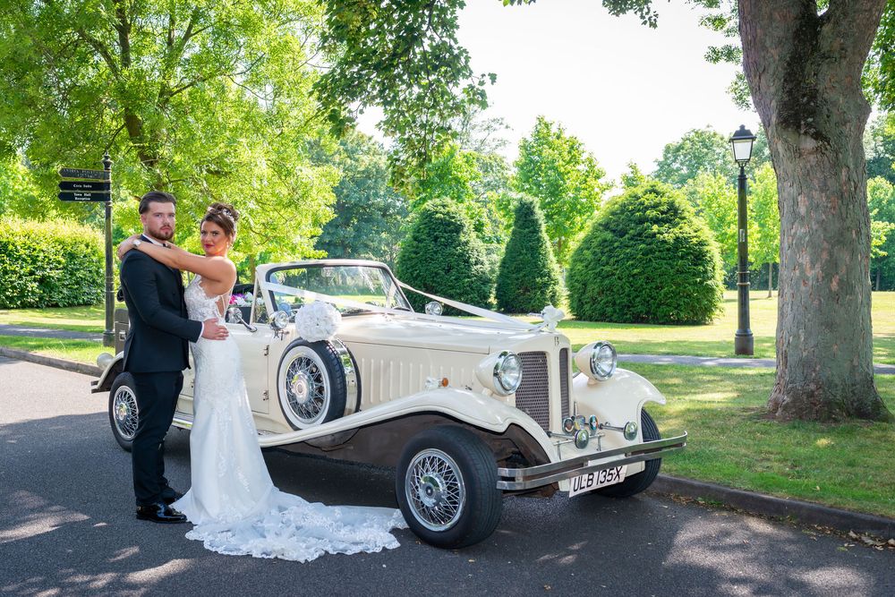 Bride and groom posing beside a classic vintage cream wedding car surrounded by trees on their wedding day.