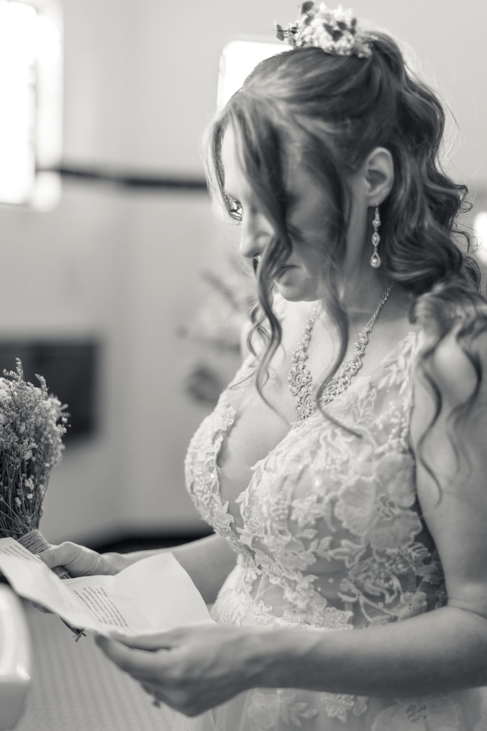 Bride in lace wedding gown reads a letter while holding flowers in a black and white photo.