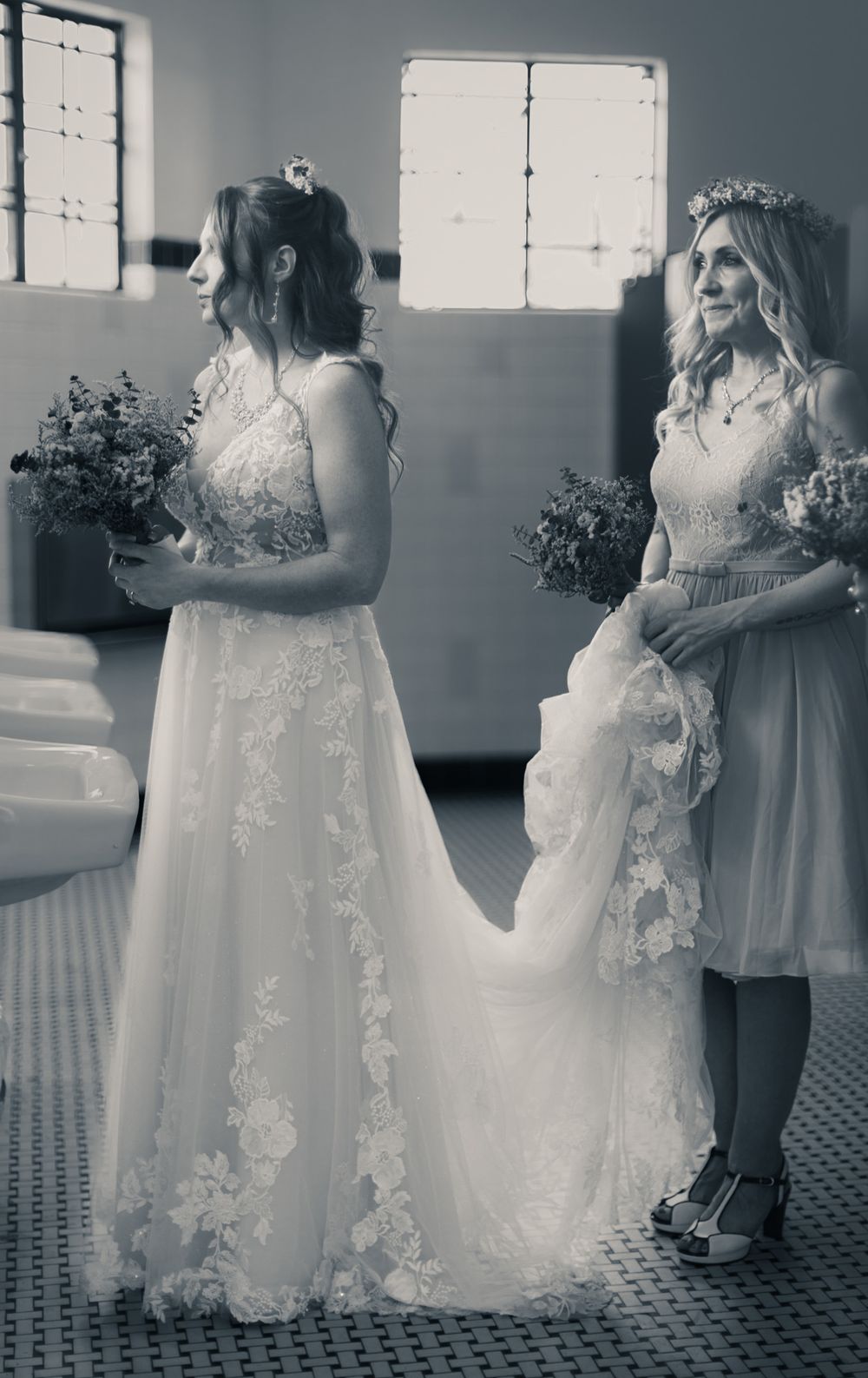 Black and white photo of a bride in lace gown holding bouquet with bridesmaid in floral crown at wedding venue.