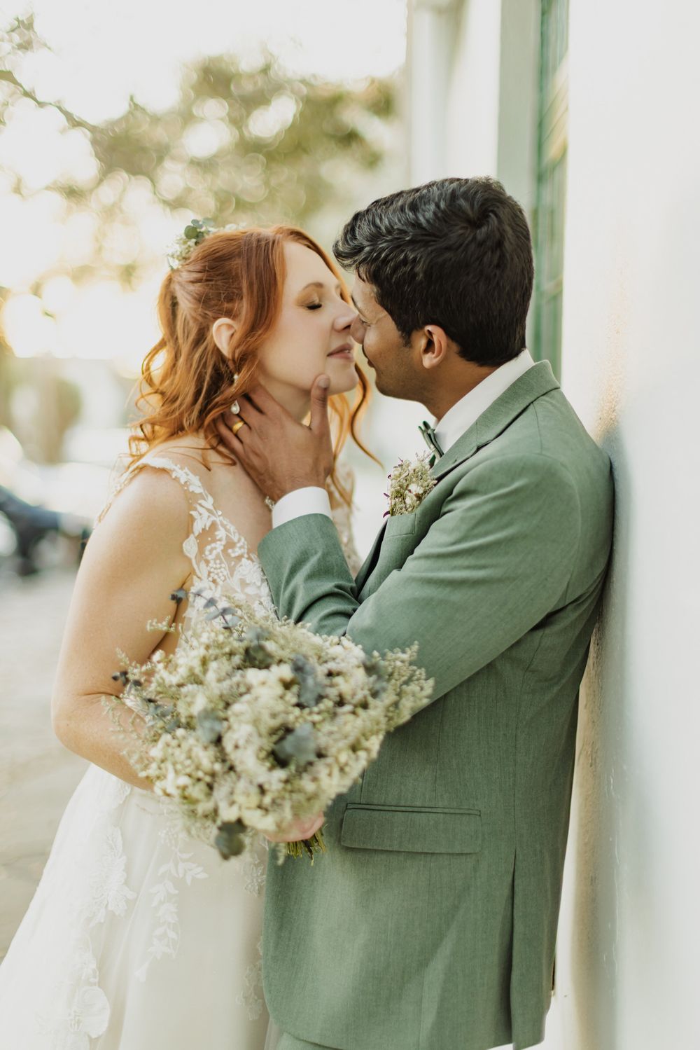 A bride and groom share a romantic kiss, she holds a white bouquet, he wears a sage green suit, leaning against a wall.