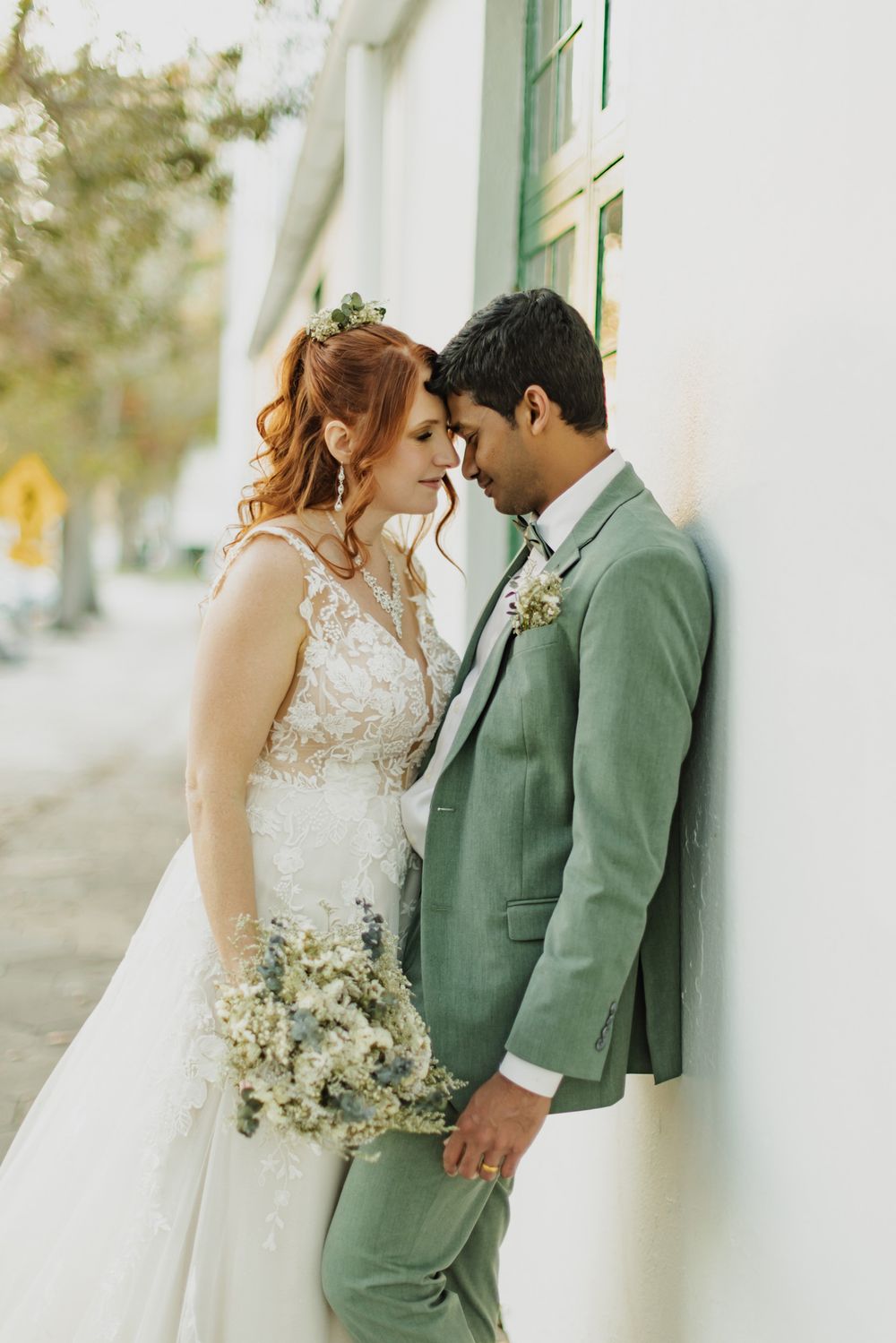 Bride and groom in sage green suit lean foreheads together against white wall, bride holding wildflower bouquet.