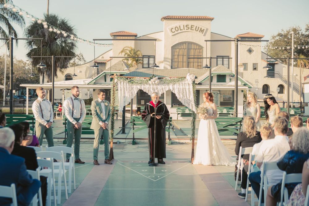 Outdoor wedding ceremony at the Coliseum venue with bride, groom, and officiant under a floral arch, guests seated in white chairs.