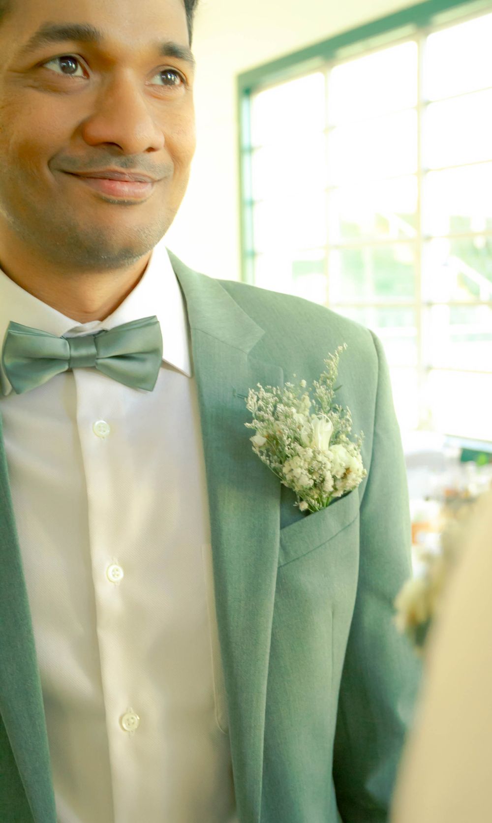 A smiling groom wearing a sage green tuxedo jacket with matching bow tie and white floral boutonniere at a wedding.