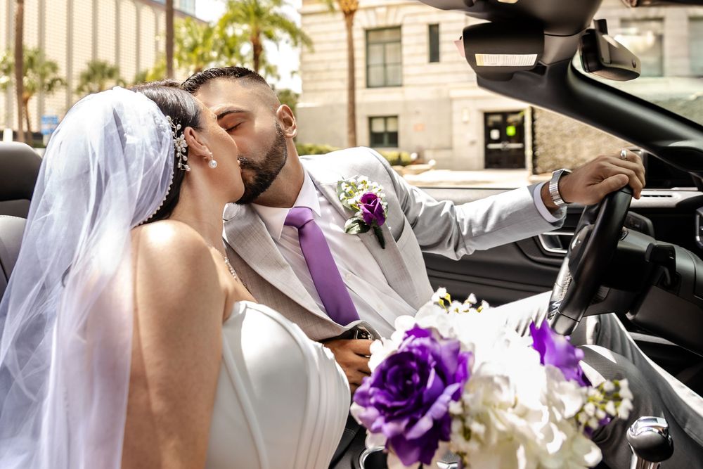 Bride and groom kissing in a convertible car, bride holding purple and white bouquet, groom in white suit with purple boutonniere.