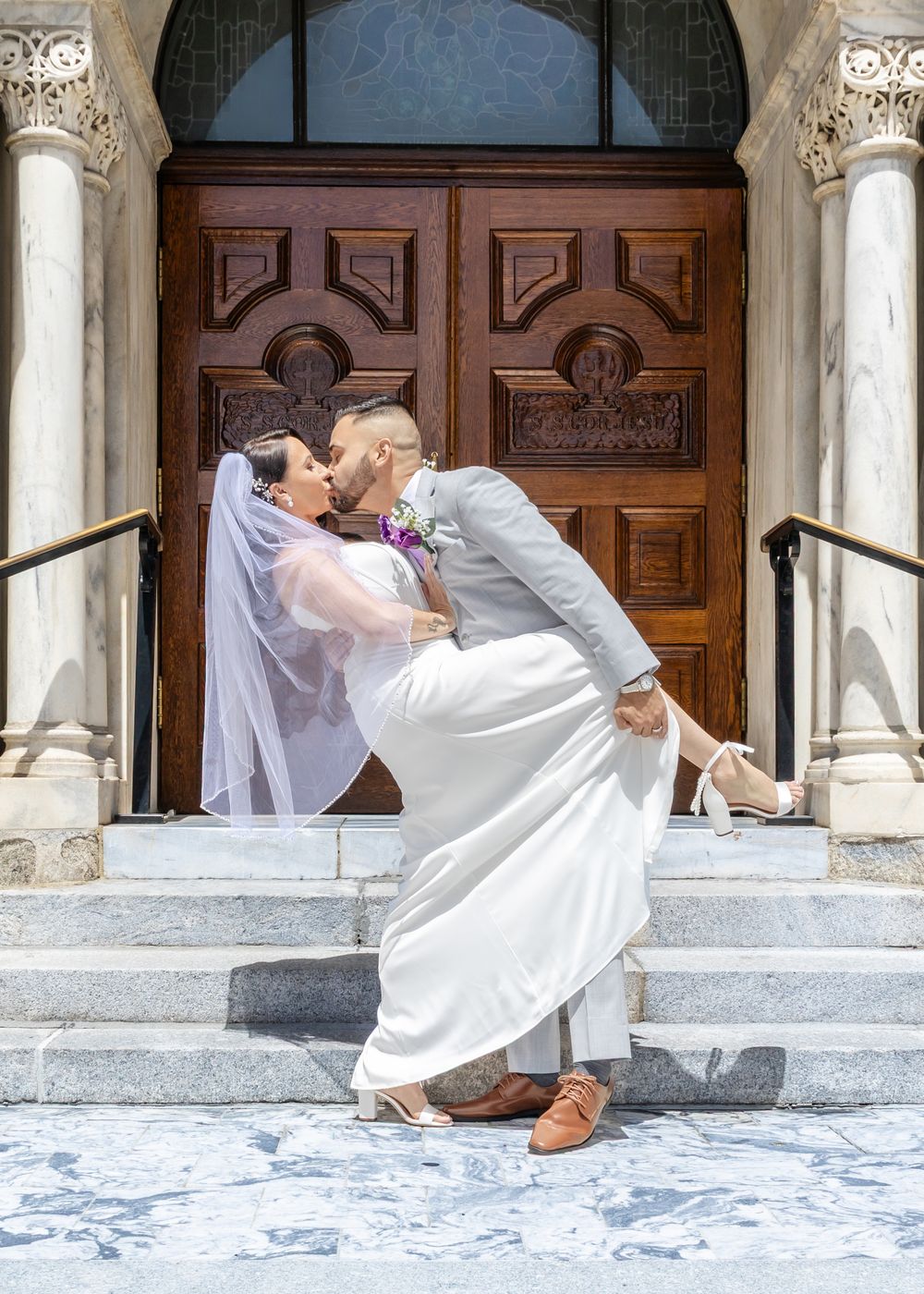 Bride and groom share a romantic dip kiss on marble steps before ornate wooden church doors on their wedding day.