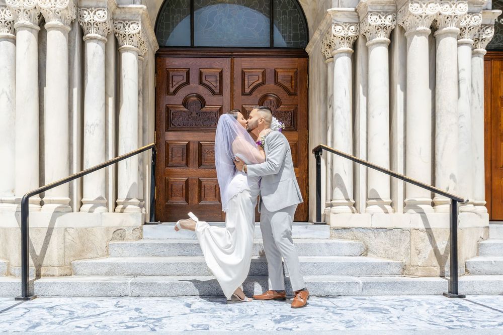 Couple sharing a romantic kiss on church steps, bride in white gown, groom in gray suit, ornate stone columns backdrop.
