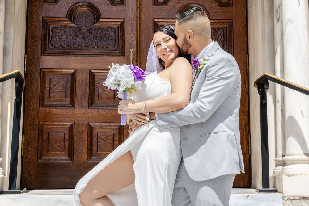 Bride and groom embrace on church steps, bride holding purple and white bouquet, groom in gray suit.