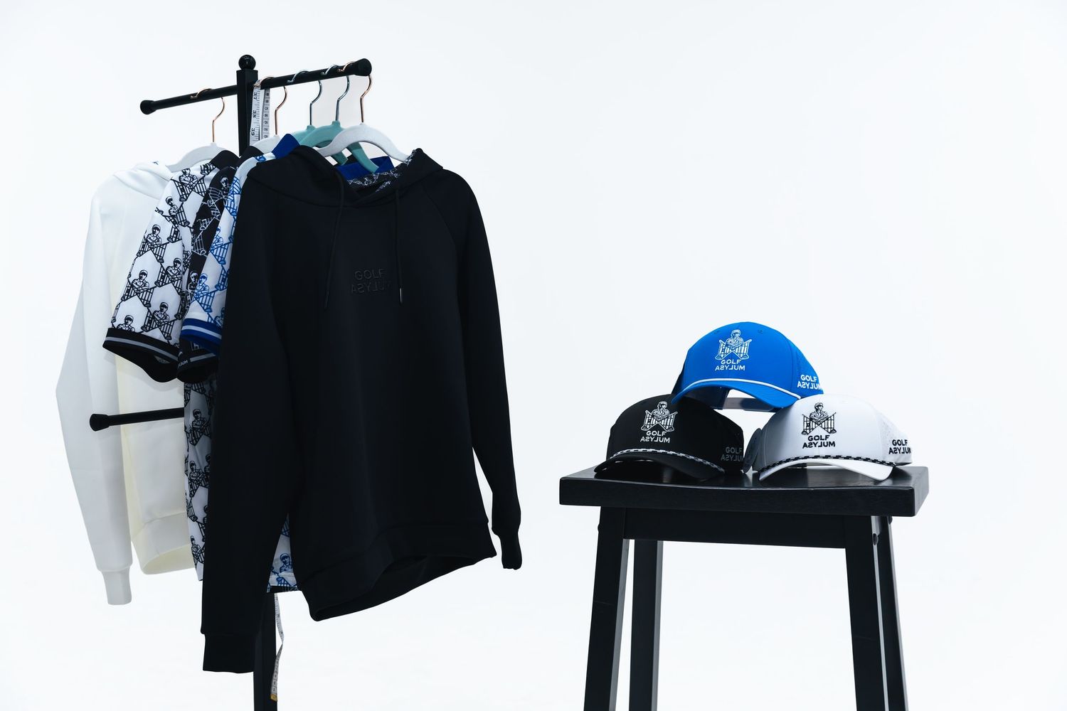 A clothing rack with black hoodie and patterned shirts beside a stool displaying blue and white snapback caps.