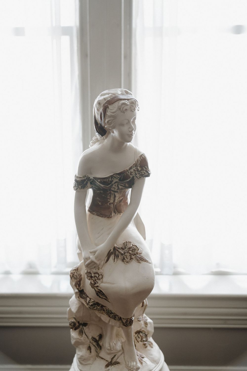 Antique porcelain figurine of an elegant Victorian woman in a floral dress, displayed near a bright window.