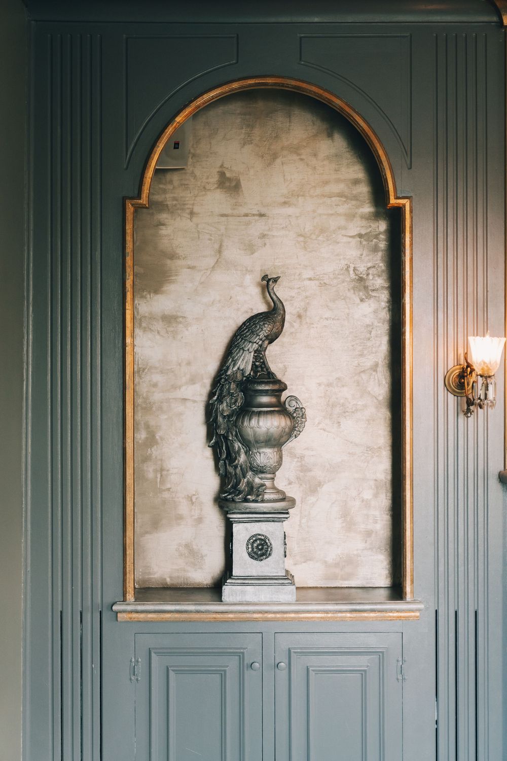 Elegant arched wall niche with gold trim featuring a bronze peacock sculpture atop a decorative urn pedestal.