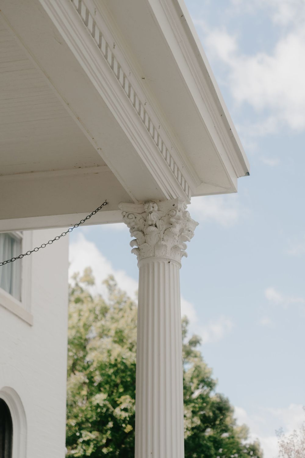 White Corinthian column with ornate capital supporting a classical porch entablature against a blue sky.