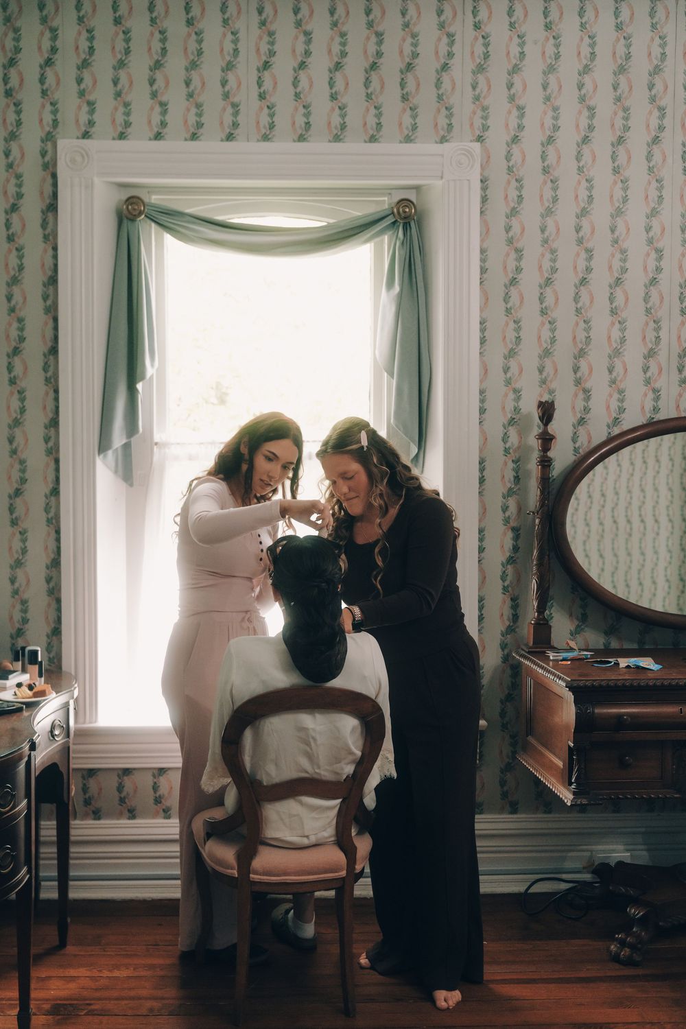 Two women helping a bride get ready near a bright window in a vintage floral wallpapered dressing room.