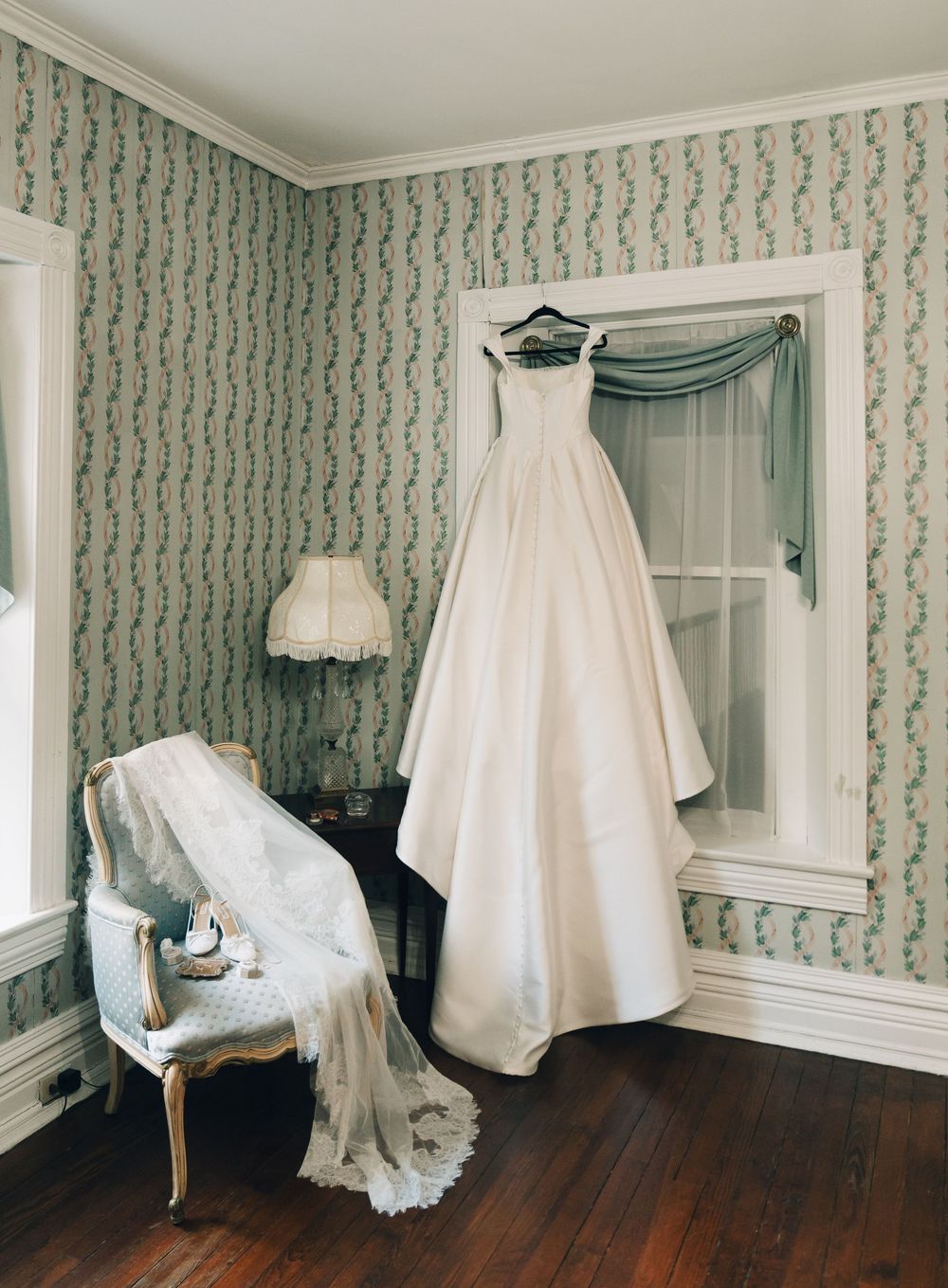 Elegant white wedding gown hanging in a vintage room with floral wallpaper, hardwood floors, and a lace-draped antique chair.