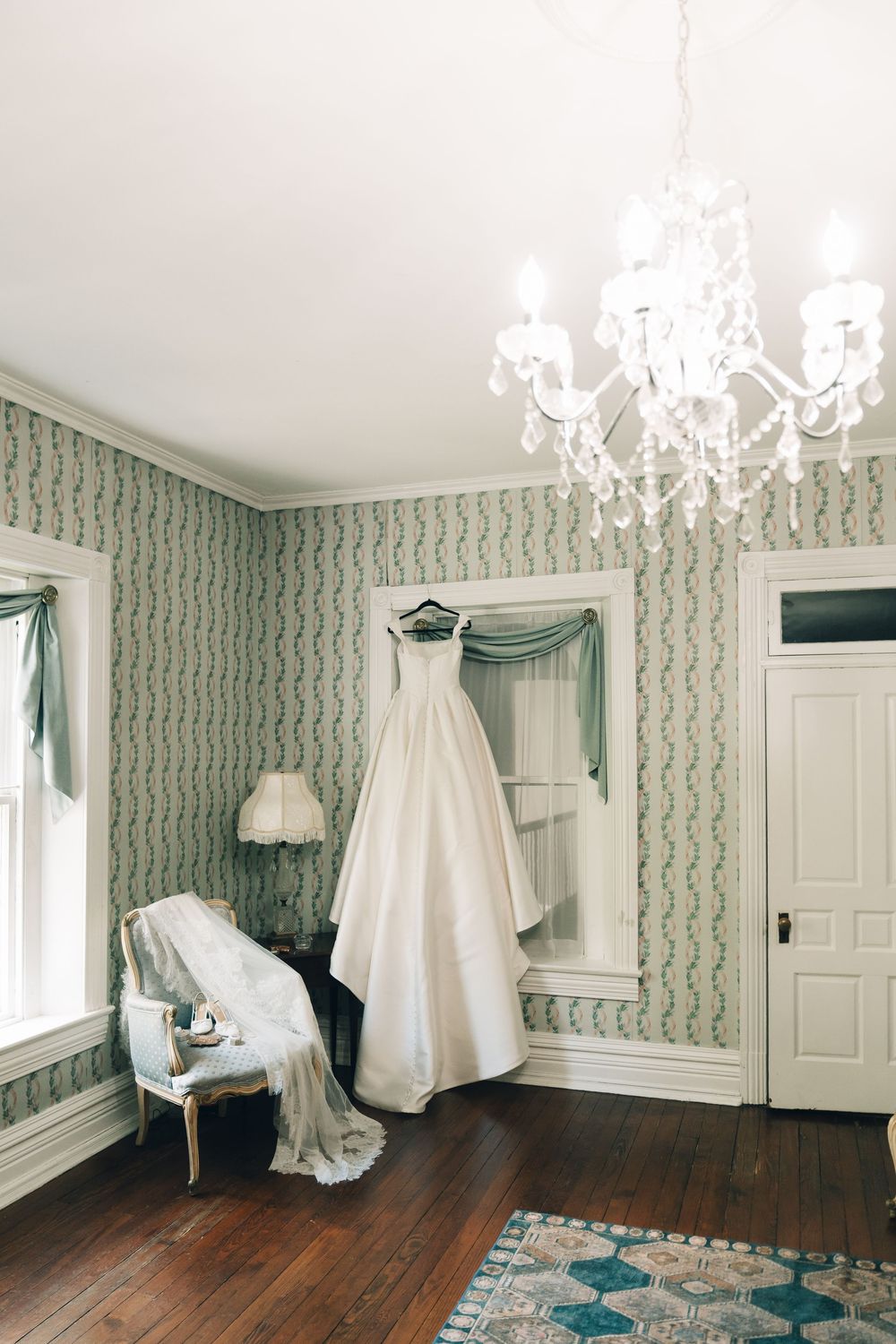 White ball gown wedding dress hanging in a vintage room with green wallpaper and crystal chandelier.