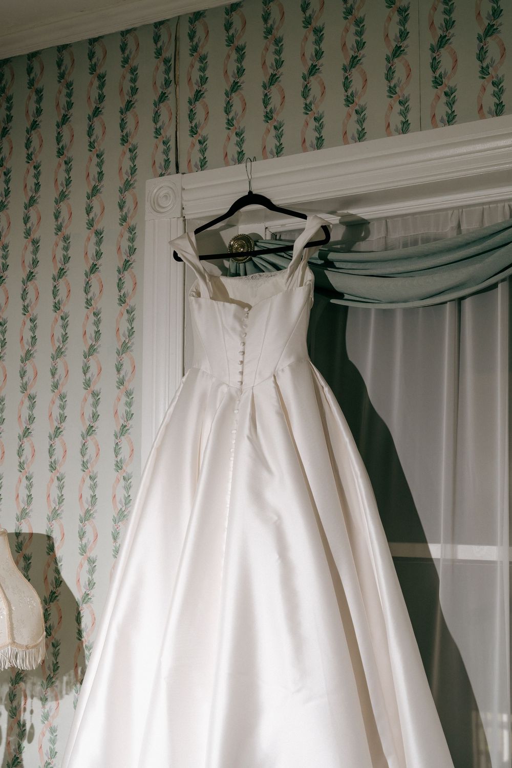 Elegant white satin ball gown wedding dress hanging on a hanger against floral wallpaper near a window.