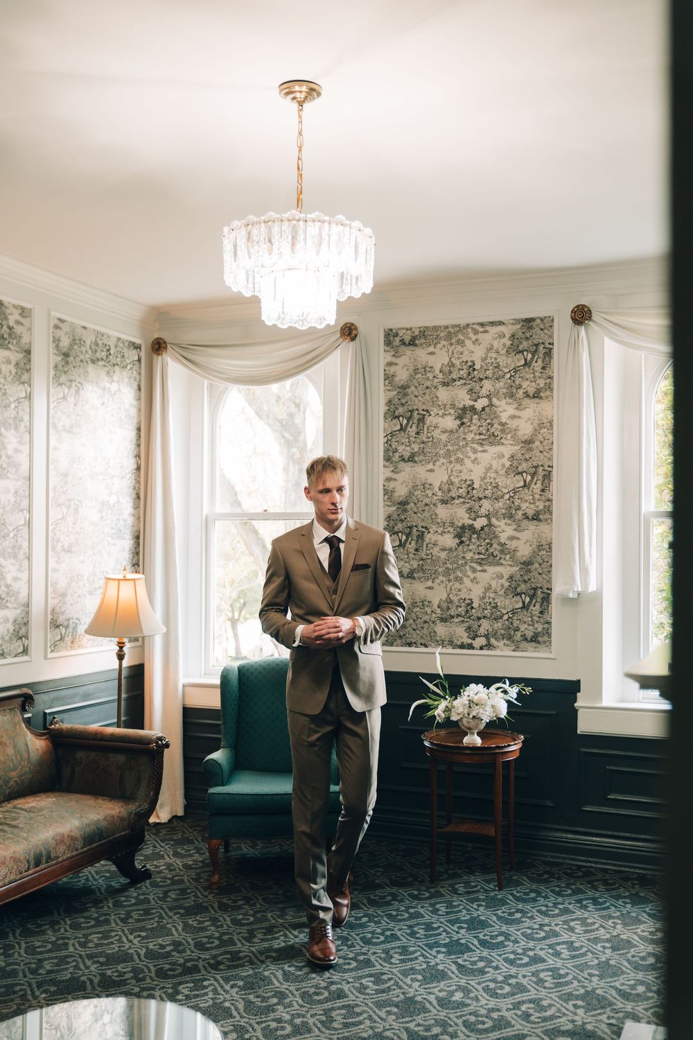 Groom in brown suit adjusting jacket in elegant room with crystal chandelier and toile wallpaper.