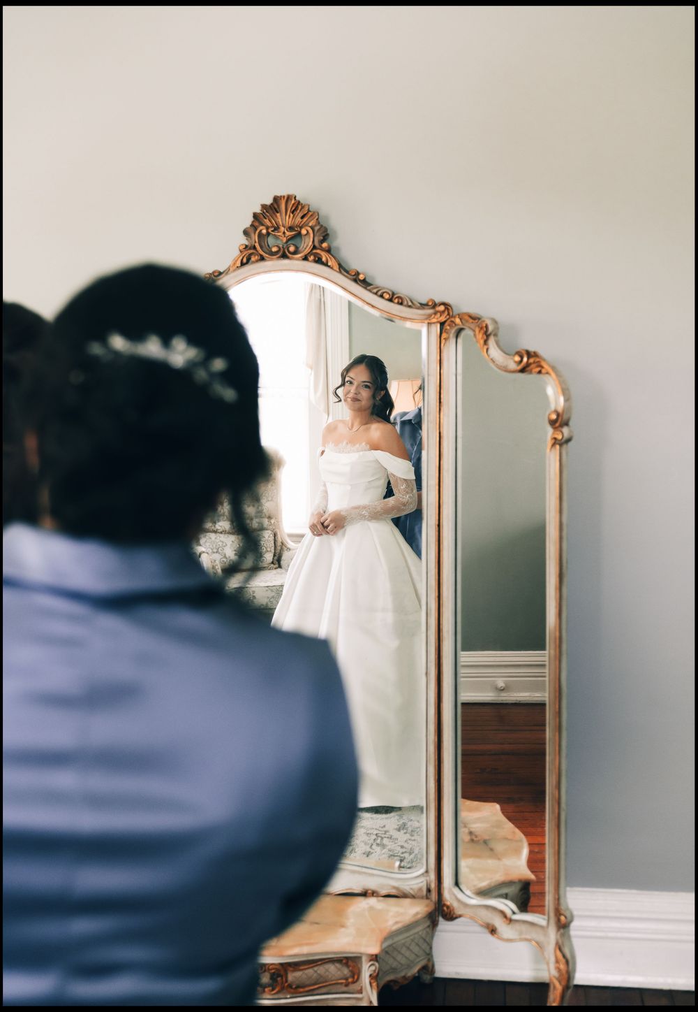 Bride in white off-shoulder gown admires her reflection in an ornate gold floor mirror during wedding preparations.