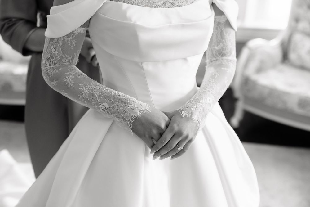 Black and white photo of a bride in a white ball gown with lace gloves, hands clasped elegantly at her waist.