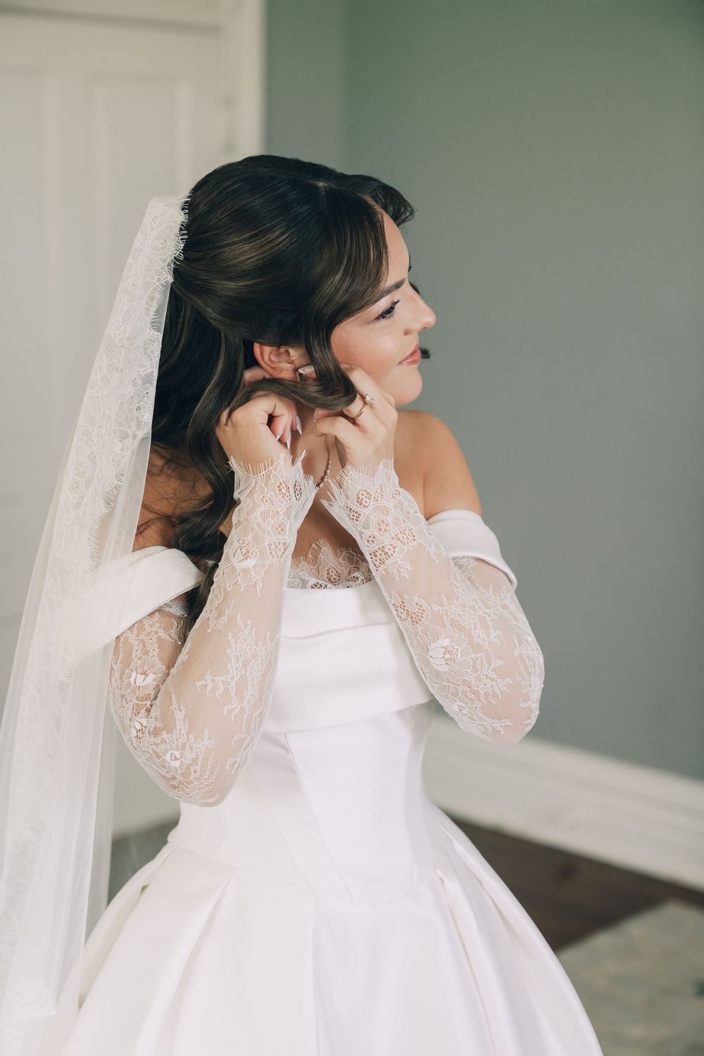 Bride in off-shoulder wedding gown with lace gloves adjusting earring while wearing a lace-trimmed veil.