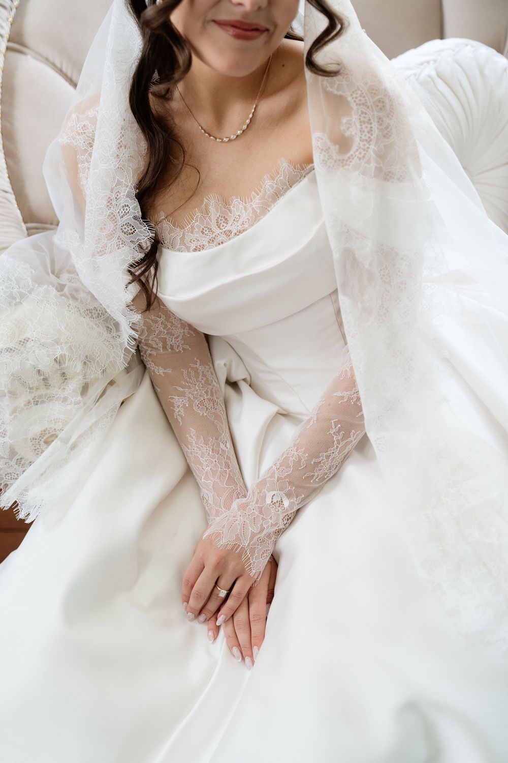 Bride in elegant white lace wedding gown with veil, pearl necklace, and diamond ring, seated gracefully.