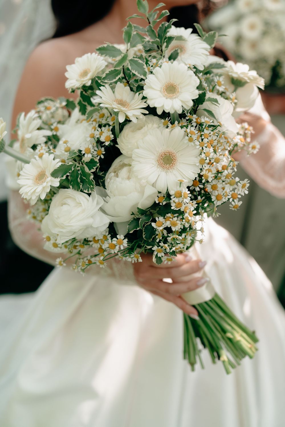 Bride holding a lush white wedding bouquet with gerbera daisies, ranunculus, and chamomile flowers.
