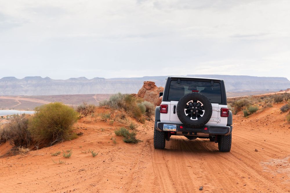 White Jeep Wrangler driving on red dirt trail with scenic desert canyon and lake views in background.