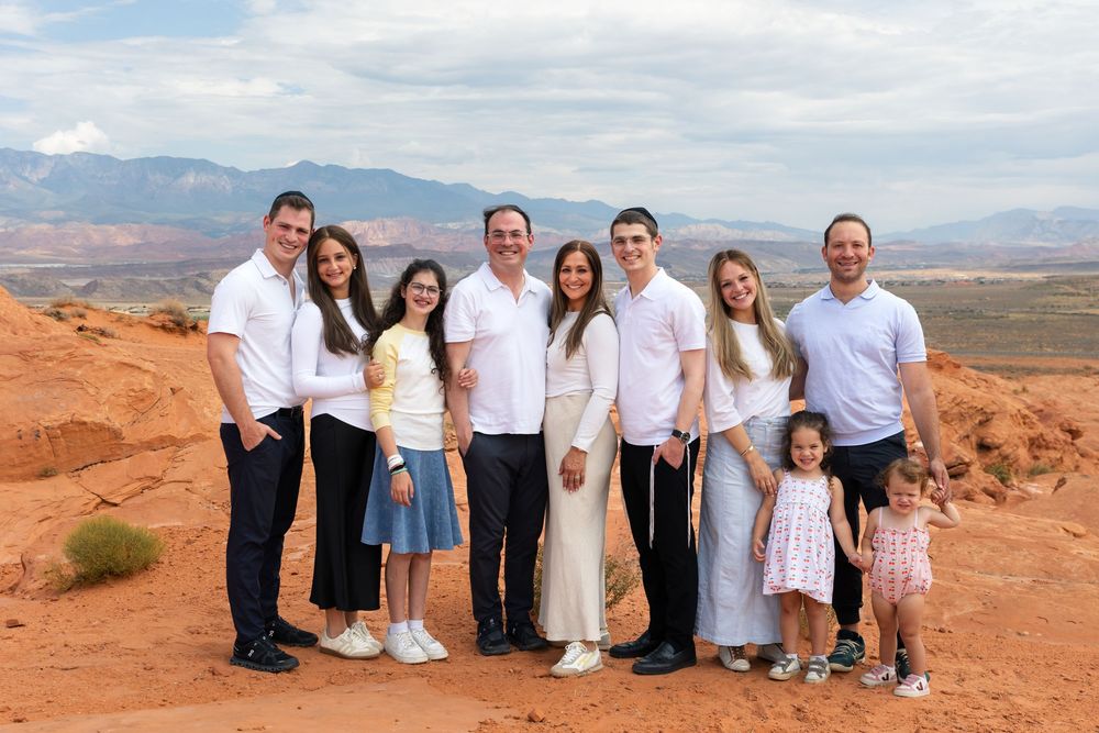 Family of nine poses together against red rock desert landscape with mountain backdrop under cloudy sky.