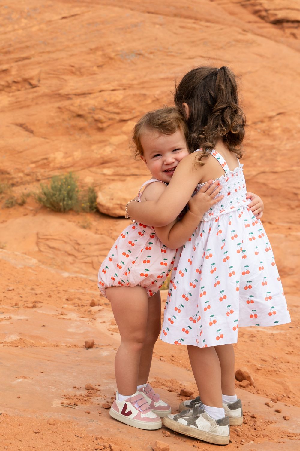 Two toddlers in cherry-print outfits hugging each other on red sandstone rock in a desert landscape.