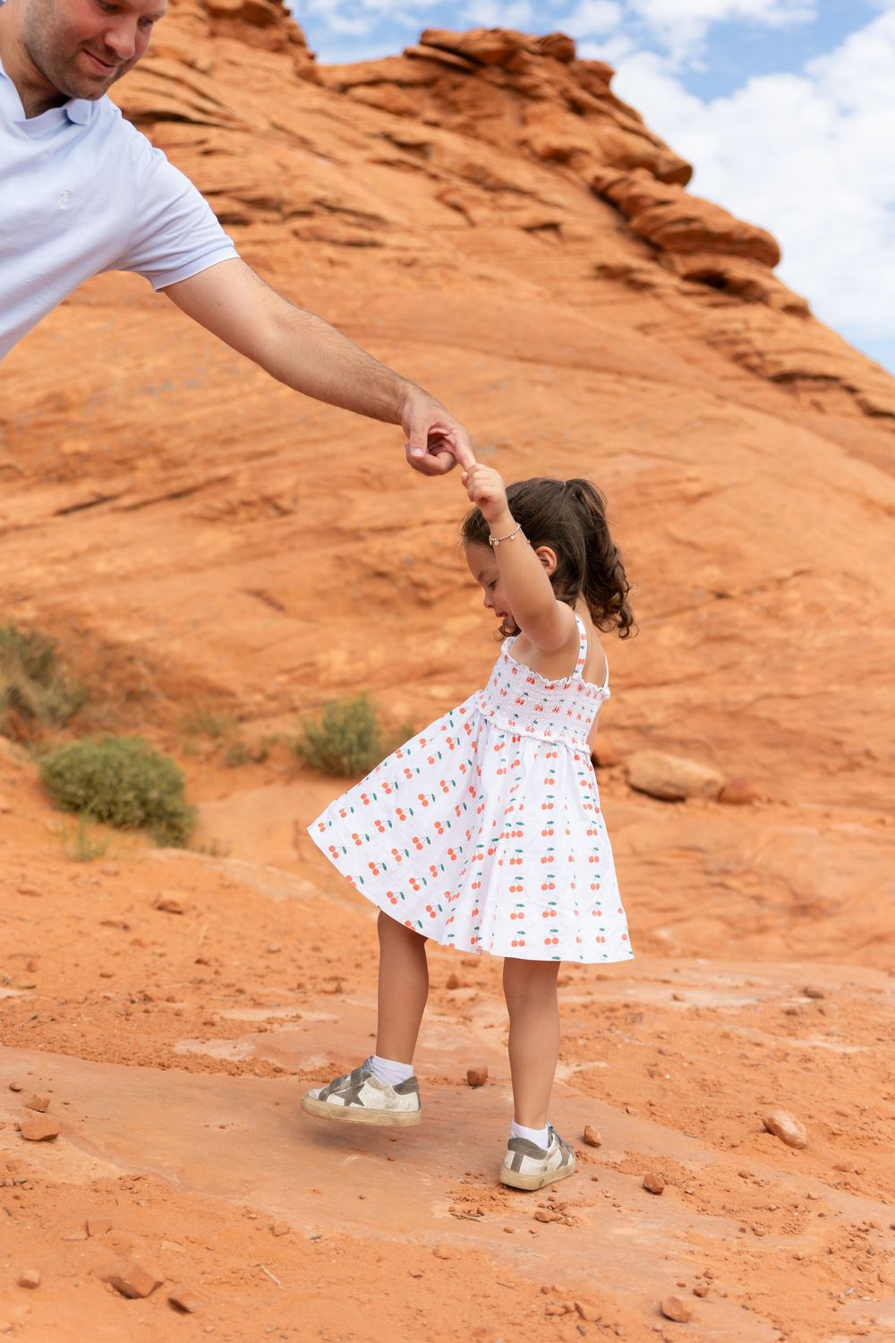 A young girl in a white cherry dress holds her father's hand while exploring red rock desert landscape.