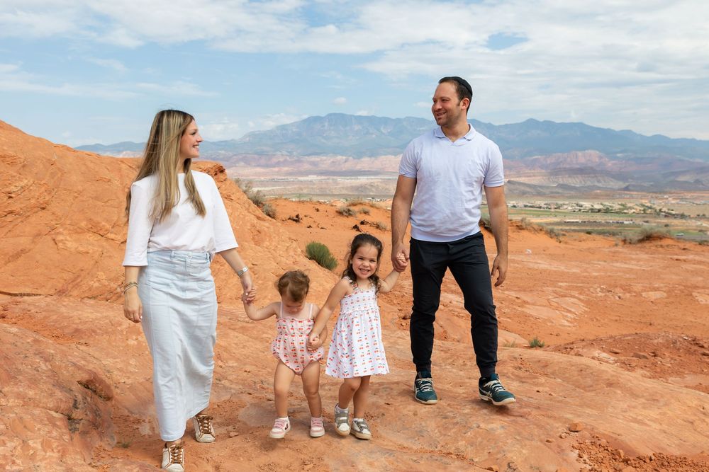 Family of four walking hand-in-hand on red rock terrain with scenic mountain backdrop under partly cloudy skies.